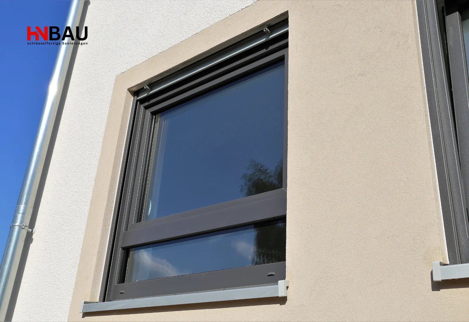 Modernes, dunkel gerahmtes Hausfenster mit integriertem Rollladen an einer hell verputzten Fassade unter blauem Himmel.