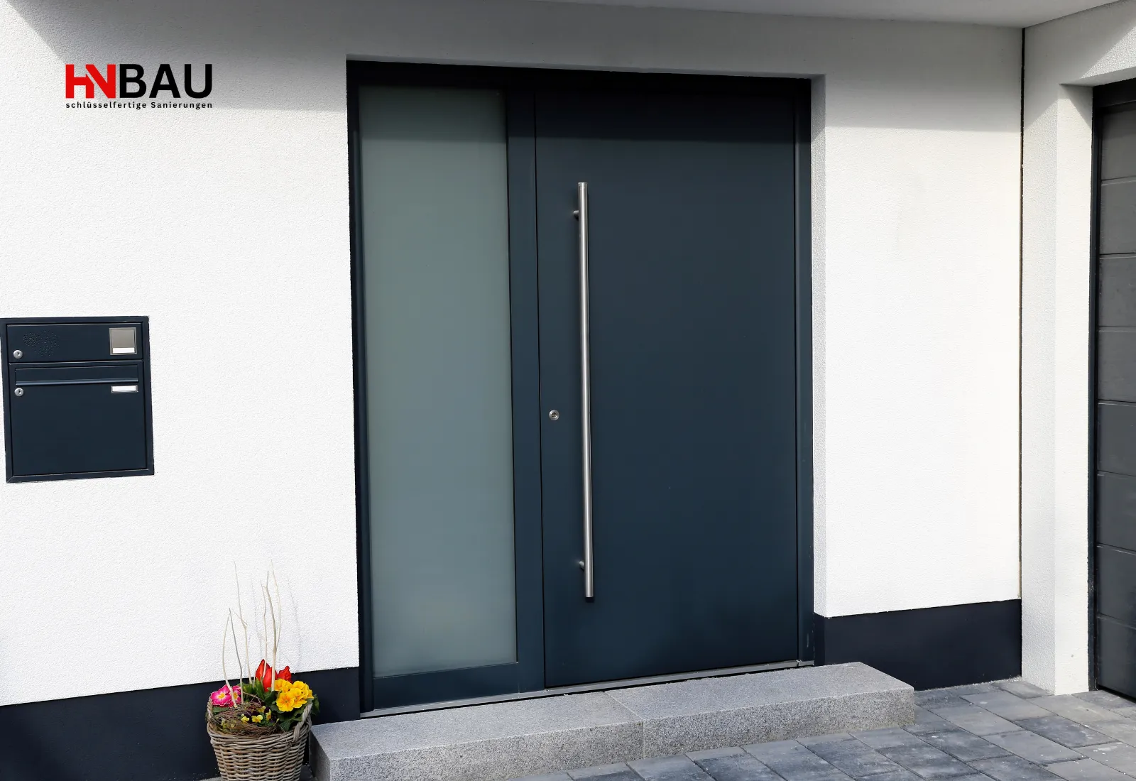 Moderne dunkelgraue Haustür mit seitlichem Glasfenster, Edelstahlgriff und weiß verputzter Fassade, daneben ein eingelassener Briefkasten und ein dekorativer Blumenkorb.