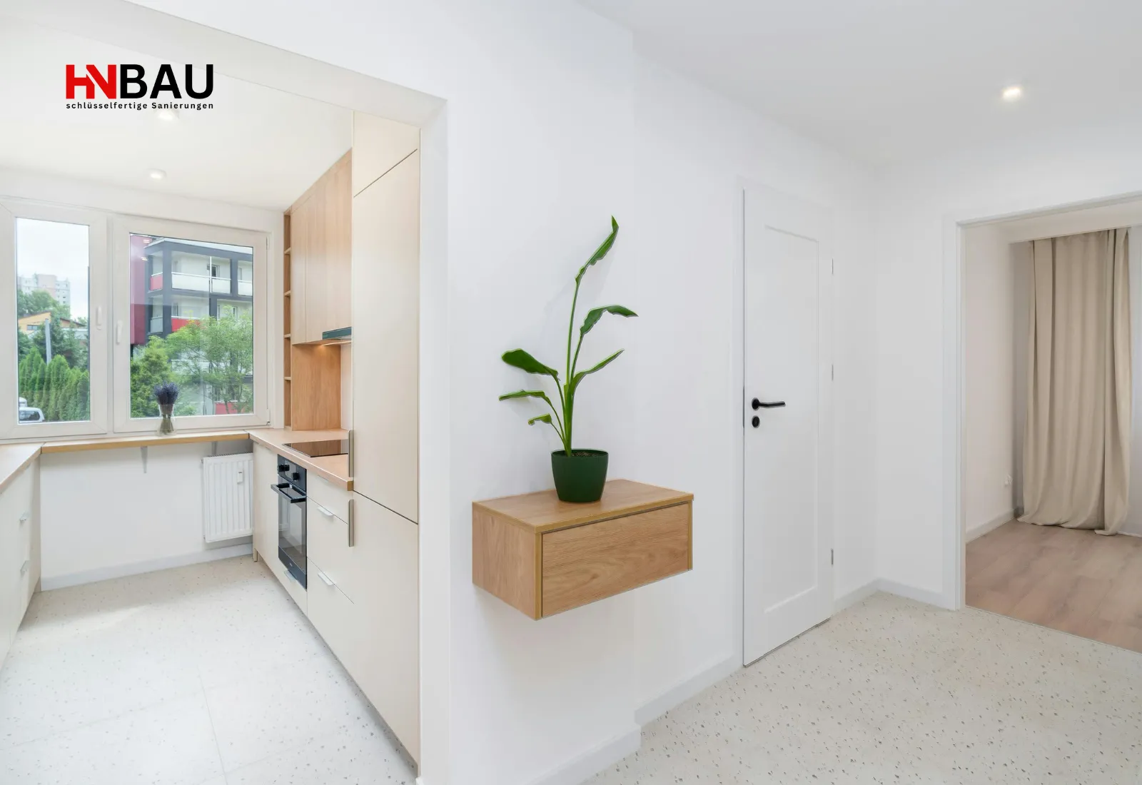 Heller, modern renovierter Wohnbereich mit weißer Innentür, offener Küche mit Holz- und Weißfronten sowie einer schwebenden Wandkonsole mit Zimmerpflanze; großes Fenster sorgt für natürlichen Lichteinfall.