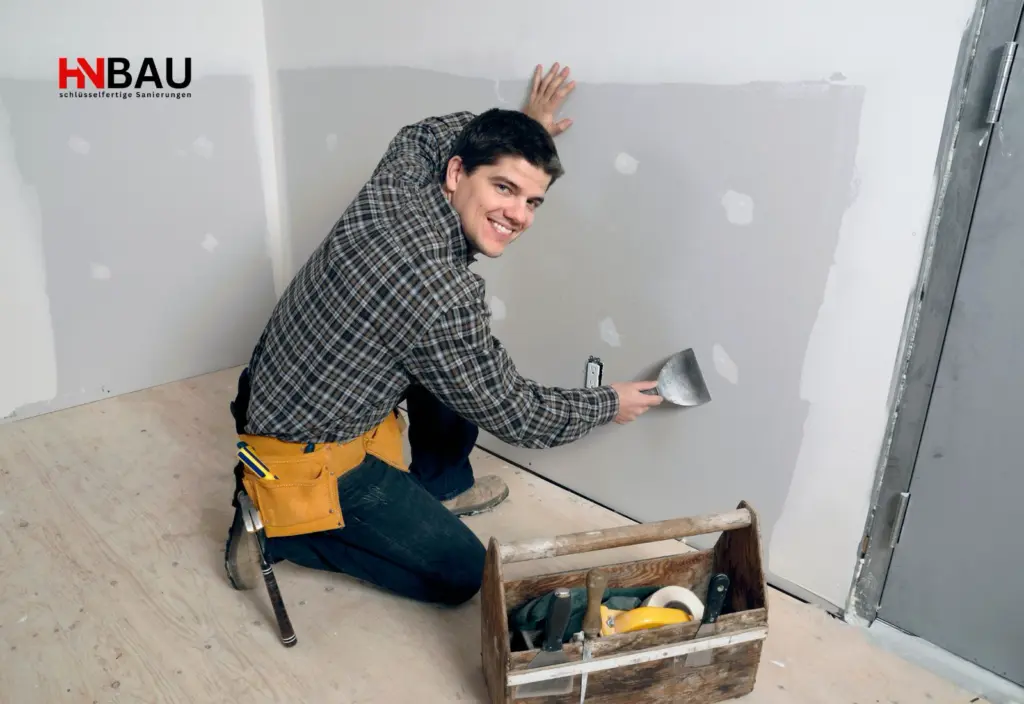 Handwerker kniet auf einer Baustelle und verspachtelt eine Innenwand; Werkzeugkiste und Arbeitsmaterial liegen daneben – Symbolbild für professionelle Renovierungsarbeiten und regionale Handwerksleistungen.