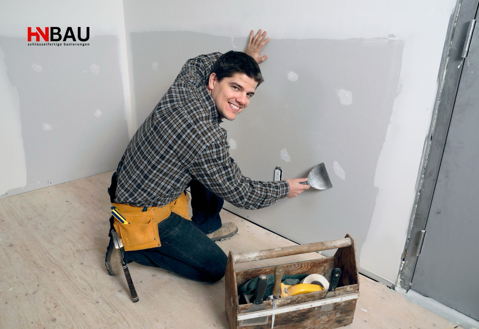 Handwerker kniet auf einer Baustelle und verspachtelt eine Innenwand; Werkzeugkiste und Arbeitsmaterial liegen daneben – Symbolbild für professionelle Renovierungsarbeiten und regionale Handwerksleistungen.