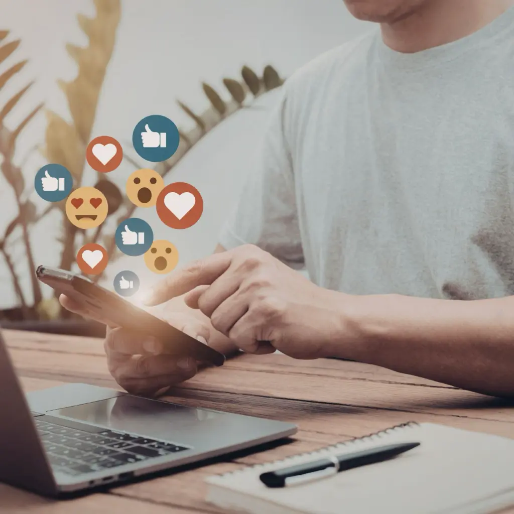 Person nutzt Smartphone am Schreibtisch mit Laptop und Notizblock, während Social-Media-Icons wie Likes, Herzen und Kommentare über dem Bildschirm erscheinen.